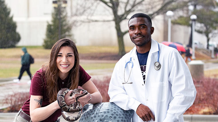 Student in a white lab coat standing next to student holding a snake outdoors.