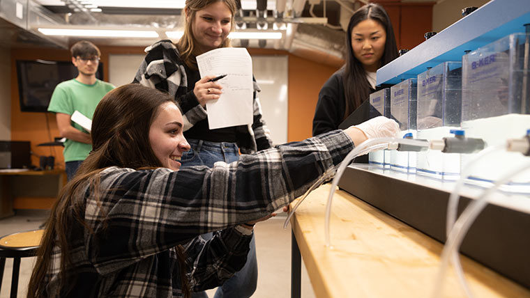 A female student using a water beaker to test the chemical properties of concrete during an engineering lab. Two other students in the background are watching her.