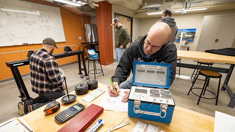 An engineering student writing down experiment results during a lab exercise. Three of his classmates are lingering around in the background.