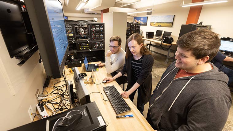 A teacher's assistant stands between two students while using a computer workstation during an electrical engineering lab.