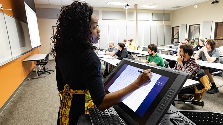 Teacher writing notes on a tablet during a lecture. Students are seated in the classroom.