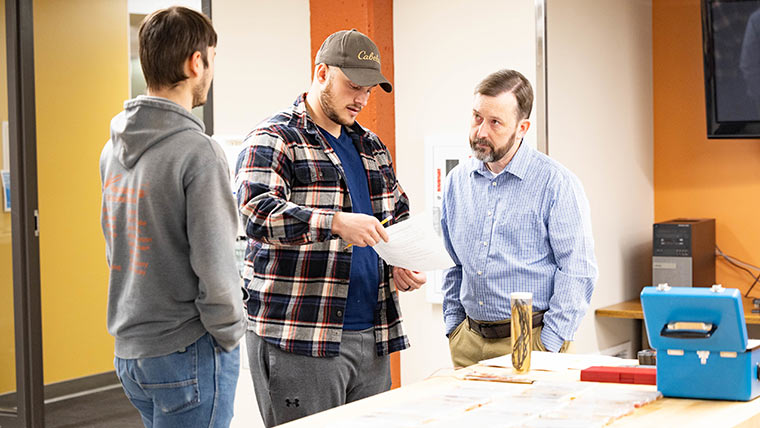 Two engineering students asking a professor a question during lab class. One of the students is holding up a piece of paper.