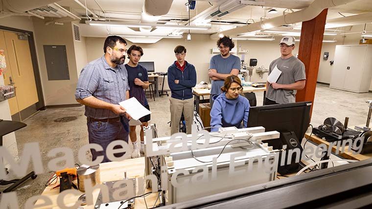 A mechanical engineering student using equipment in a lab while a professor and classmate watch him.