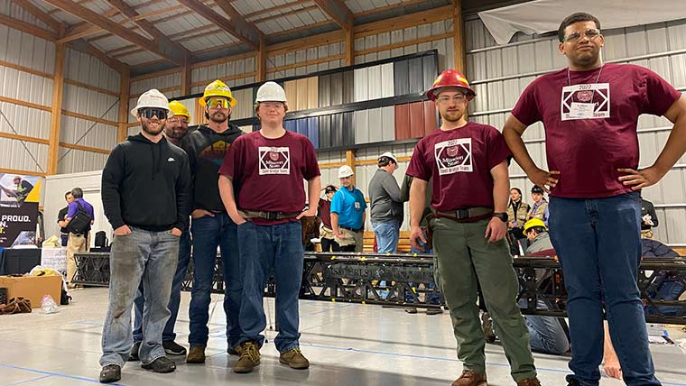 Missouri State Steel Bridge team posing for a photo at a competition event. In the background is the steel bridge they made.