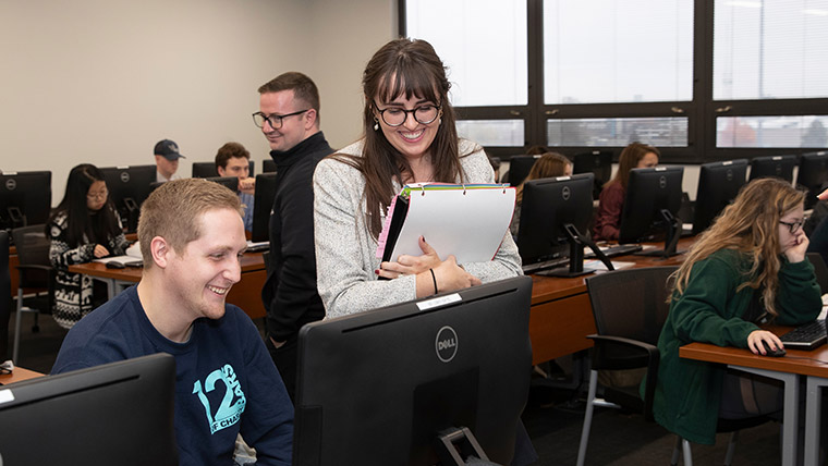 Teacher looking over the shoulder of a student within a classroom of computers.