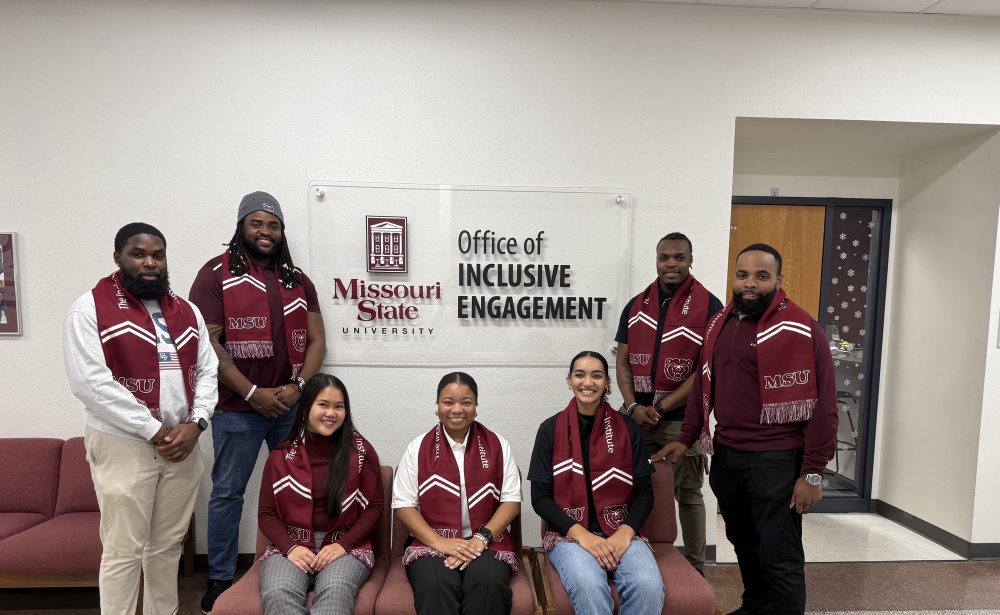 7 GA's standing outside of the Office of Inclusive Engagement wearing maroon and white "Inclusive Excellence" scarves.