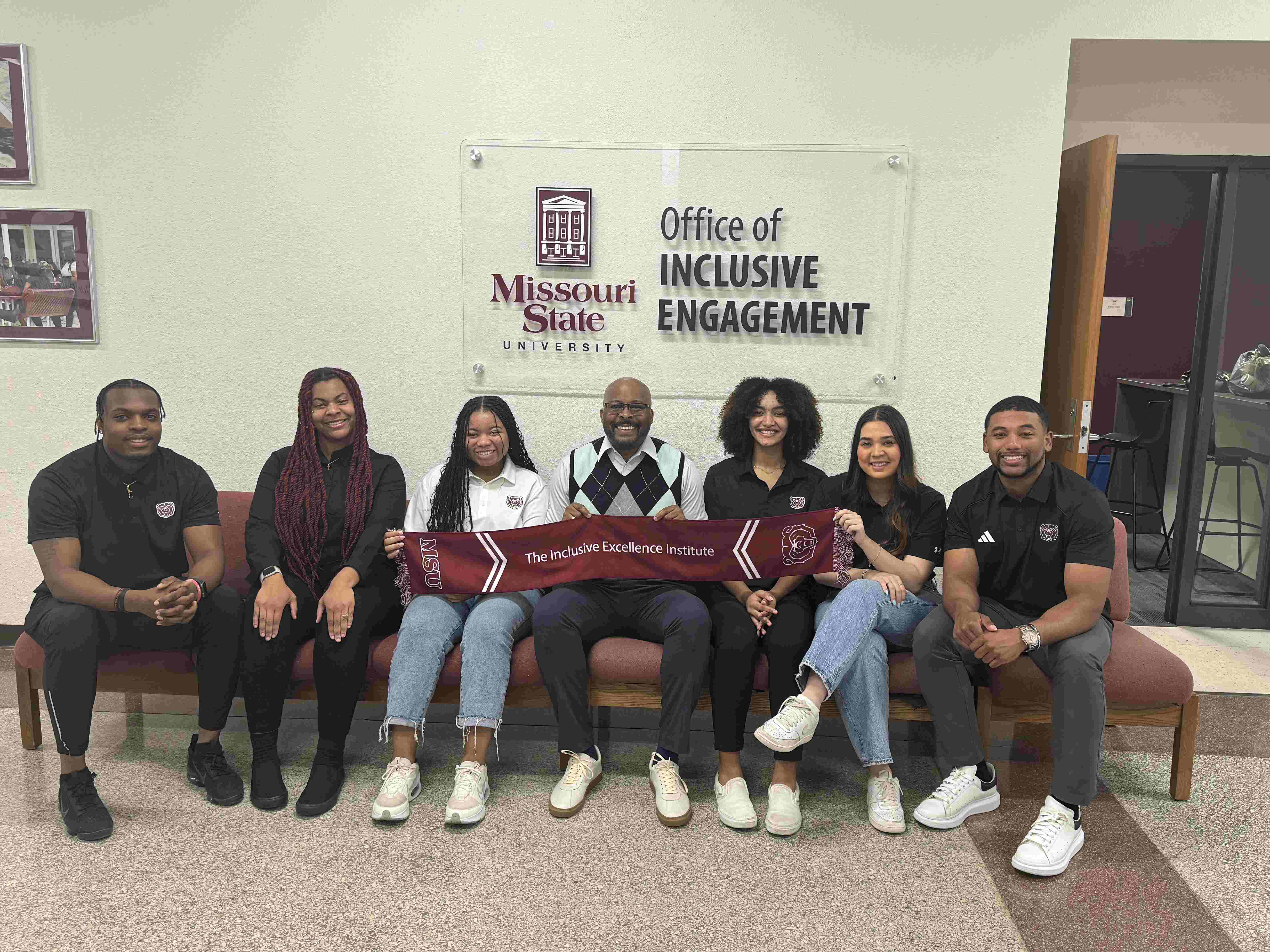 Six graduate assistants from the Office of Inclusive Engagement sitting down smiling with the Assistant to the President for Inclusive Engagement