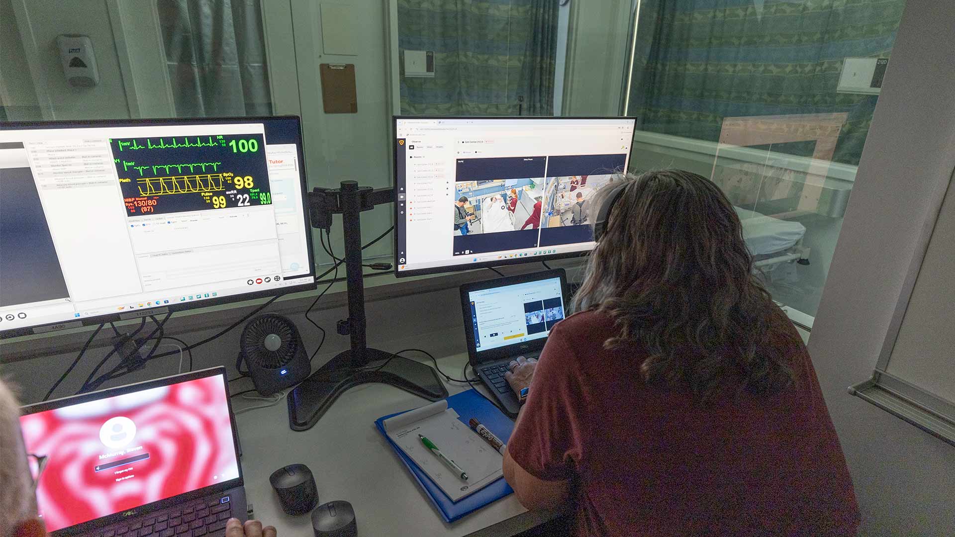 A faculty member in the simulation lab observes students training in the skills lab.