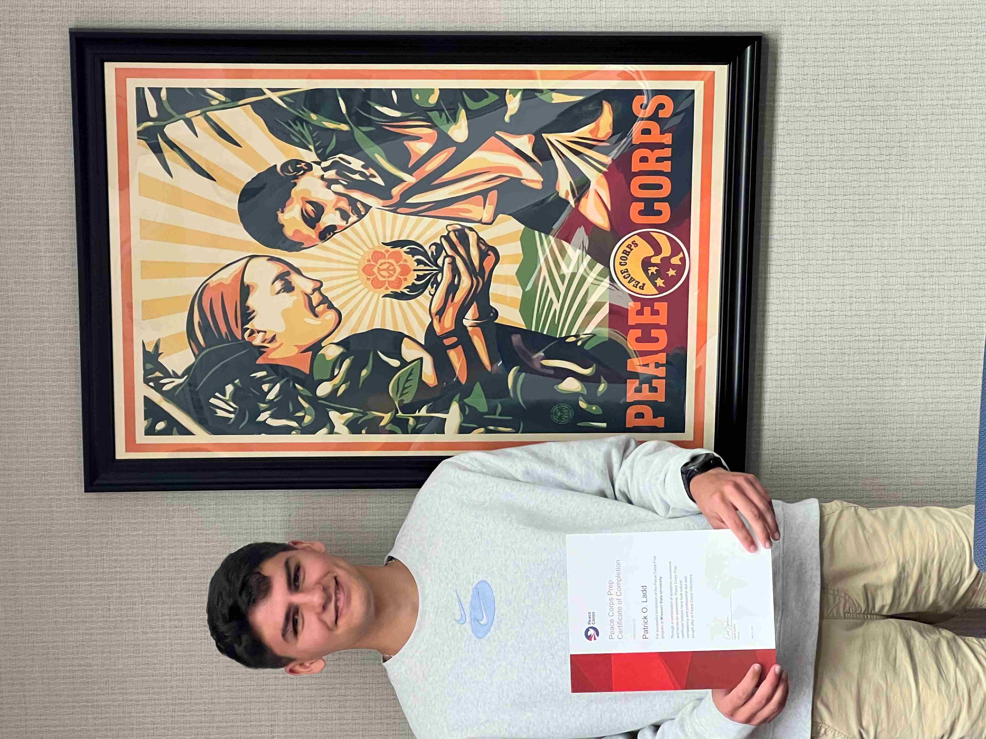 Student holding a certificate stands in front of a Peace Corps poster