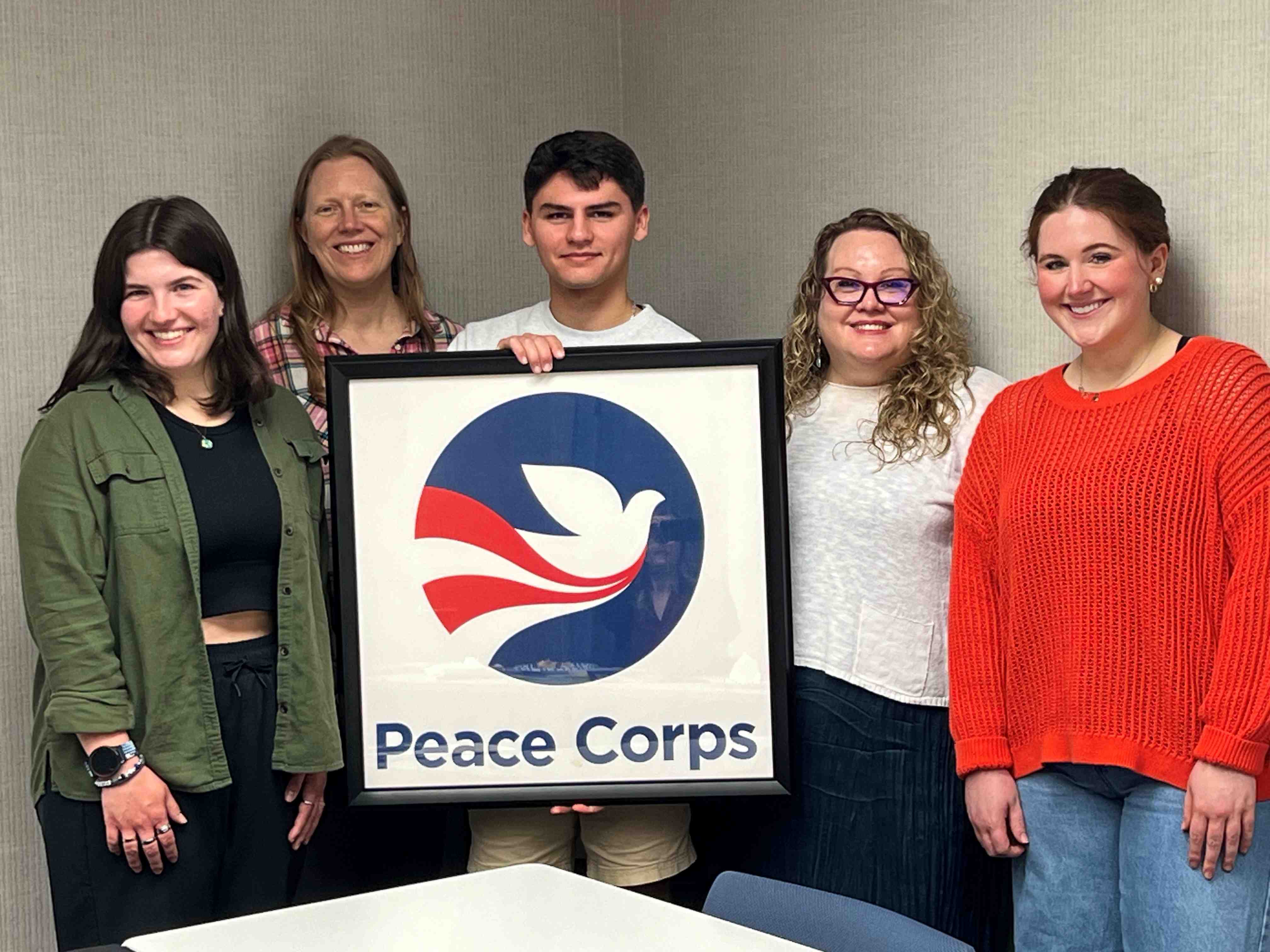 5 people stand behind a framed print of the Peace Corps logo
