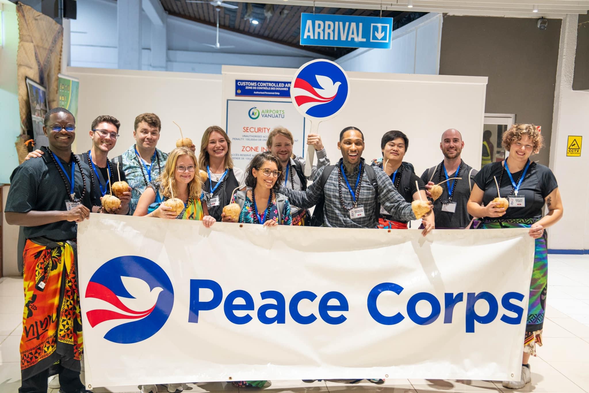 11 people stand behind a Peace Corps banner, many holding coconuts with straws for drinking from them