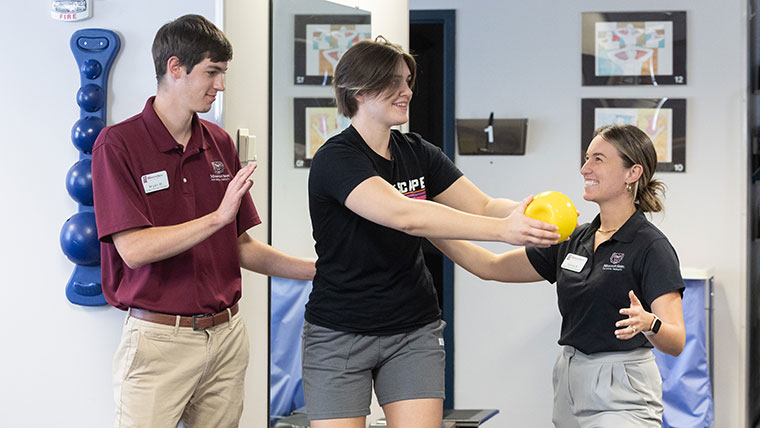 Two Missouri State physical therapy students guide a teenager through a rehab activity. The teenager is moving a medicine ball from side to side.