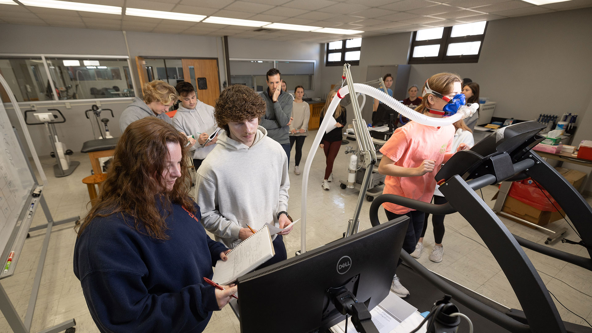 A kinesiology student keeps pace on a treadmill during a VO2 max test as two other students monitor her activity on a computer. The rest of the class discuss their notes and watch nearby.