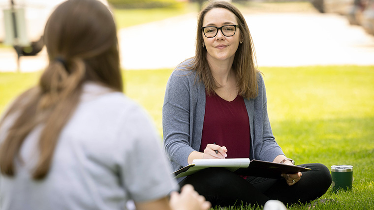 Two individuals sitting on the lawn. One individual has a clipboard and is listening to the other individual. 