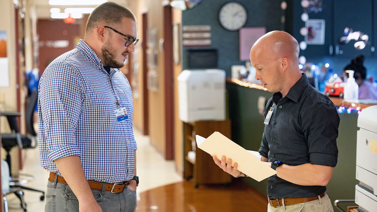 Two individuals within a hospital. One is reviewing the contents of a folder while the other individual waits. 