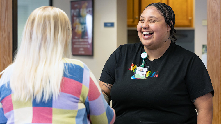 Two individuals within a clinic. One individual is wearing a name badge and is smiling at the other individual. 