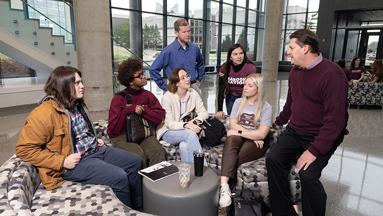 A group of students seated on a couch listen to one of their professors in Glass Hall.