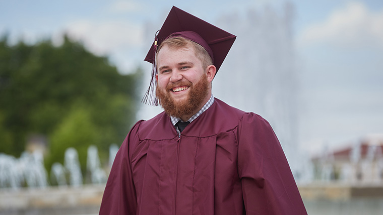 Smiling Missouri State graduate in cap and gown standing in front of John Q Hammons Fountains