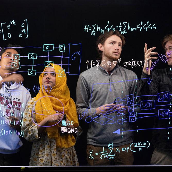 Four students study equations on a transparent markerboard for a quantum circuit design project.